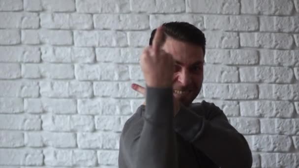 Молодая брюнетка в серой футболке на стене из белого кирпича показывает непристойный жест, средний палец, антисоциальное поведение — стоковое видео