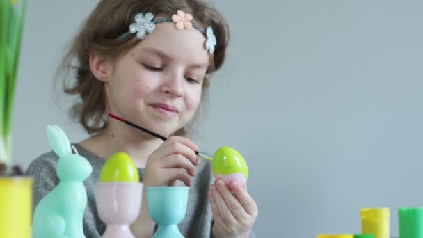 Teenie-Mädchen bereitet Dekorationen für das Osterfest vor. Ostereier mit Pinsel bemalt, verlegen lächelt — Stockvideo