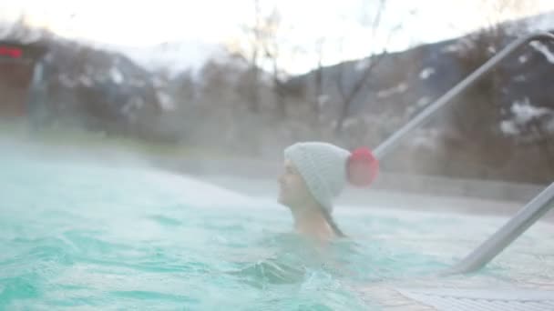 Belle brune en bonnet gris tricoté avec un pompon rouge et un maillot de bain nage dans une source thermale sur fond de montagnes enneigées en hiver — Video