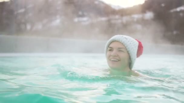 Женщина в шляпе и купальнике плавает в горячем открытом бассейне зимой. Горячий источник в горах, зимние каникулы, санаторий — стоковое видео