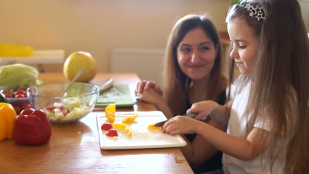 Loving mother kisses her daughter. A girl learns to cut vegetables for salad in the kitchen, her mother helps her — Stock Video