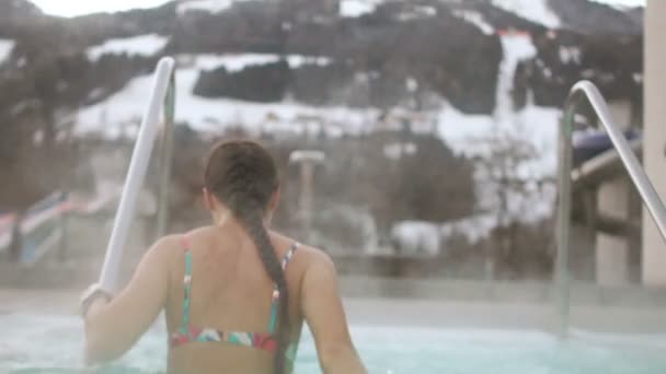 Струнка брюнетка в купальнику виходить з гарячої весни взимку на тлі засніжених гір. Від дівчаток тіло приходить пара. Відпочинок в курортному комплексі — стокове відео