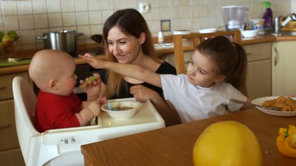 Mujer joven ama de casa alimentando a dos niños en la cocina. Niño pequeño y niña preescolar — Vídeo de stock