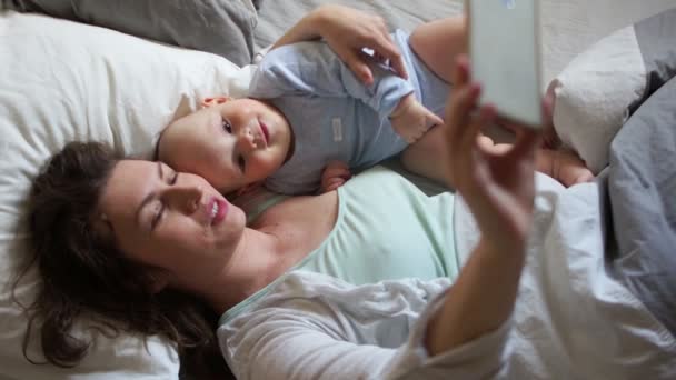 Mamá y el bebé están acostados en la cama y tomando selfies. Día de las madres, buena familia, teléfono inteligente y bebé — Vídeo de stock