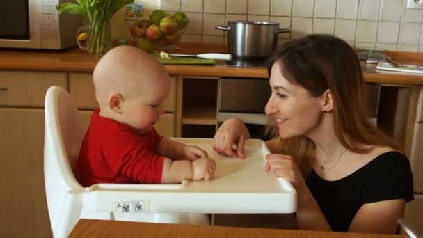 Matka, karmienie głodnych dzieci w krzesełko do karmienia w kuchni. Kobieta uśmiecha się delikatnie do dziecka. Dzień matki — Wideo stockowe