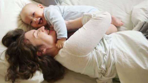 Familia soleada mañana mamá y el bebé en la cama. Una mujer hace cosquillas y abraza a su hijo de un año. Día de las Madres — Vídeo de stock