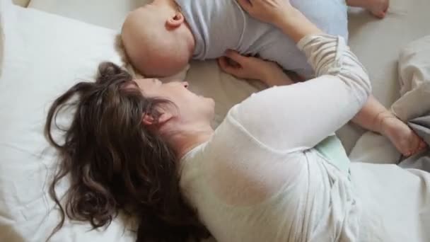 Mãe e bebê estão brincando na cama pela manhã. O bebê rola e rasteja, a mãe o devolve, o bebê ri — Vídeo de Stock