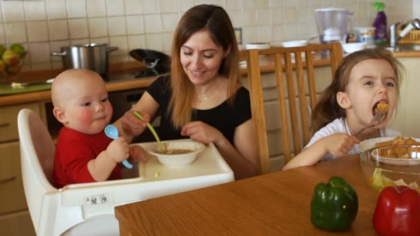 La ama de casa inteligente alimenta a sus dos hijos en la cocina. El niño está sentado en la silla y trepado en el plato con las manos. Niña preescolar come por su cuenta — Vídeos de Stock