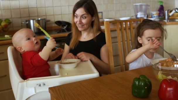 Женщина кормит двоих детей на кухне. Ребенок в кресле играет ложками и не хочет есть. Медсестра забирает у него грязную ложку. Национальное агентство персонала — стоковое видео