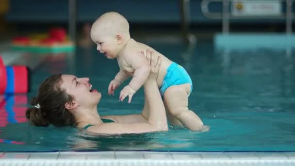 Una mujer en una piscina cubierta vomita a un bebé de un año bien alimentado. El niño se ríe alegremente. Día de las madres estilo de vida saludable — Vídeos de Stock