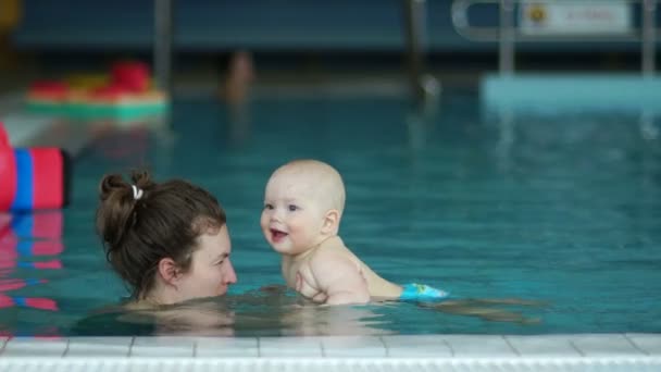 Bebé y mamá se divierten nadando en la piscina cubierta. El bebé se ríe alegremente, sus dientes de leche son visibles — Vídeos de Stock