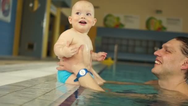 Boldog baba apjával a medencében. A gyerek ül az oldalán, és megpróbálja ugrani a vízbe, az ember támogatja őt. Egészséges életmód, a gyerekek fejlesztése — Stock videók