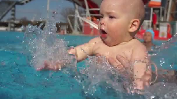 Één jaar oude baby leert om te zwemmen in het buitenzwembad met de hulp van zijn vader. Close-up, vliegen spray, versterking van de immuniteit van het kind — Stockvideo