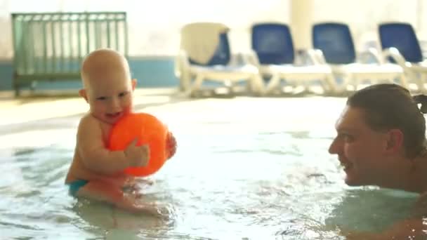 Pai carinhoso e seu bebê estão brincando com uma bola enquanto estão sentados na água em uma piscina quente para crianças. O desenvolvimento das crianças, dia dos pais — Vídeo de Stock