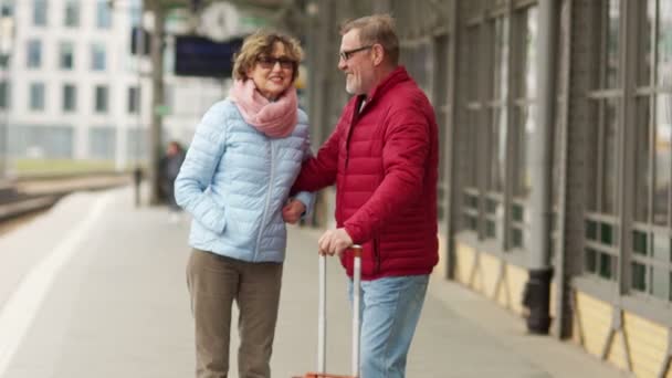 Pareja madura con una maleta en la plataforma del tren riendo y abrazándose. Jubilados de viaje. Un hombre y una mujer llevan chaquetas de primavera azules y rojas. — Vídeo de stock
