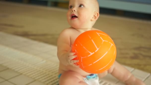 Precioso bebé está jugando con una bola naranja mientras está sentado en una piscina de niños calientes en un parque acuático — Vídeos de Stock