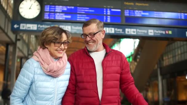Чоловік і дружина стоять на платформі залізничного вокзалу, вони сміються весело, чоловік жартома кашляє, пара тримає в руках паспорти і квитки — стокове відео