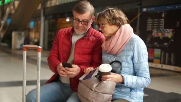 Un par de personas maduras consultan el smartphone esperando el tren. Esposo y esposa esperando el tren se sientan en un banco en la estación de tren — Vídeos de Stock