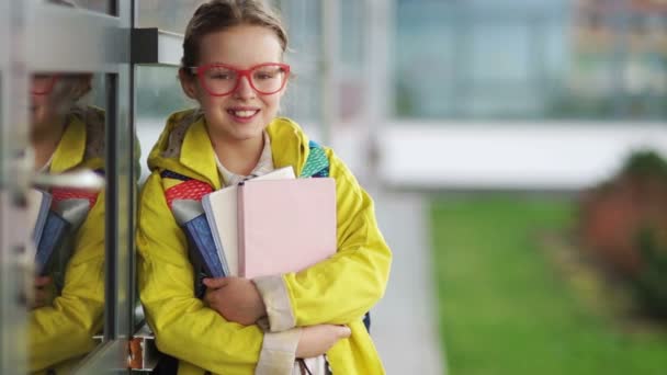 Studentessa con una giacca gialla e occhiali rossi sorride timidamente, guardando nella fotocamera. Ritorno a scuola — Video Stock