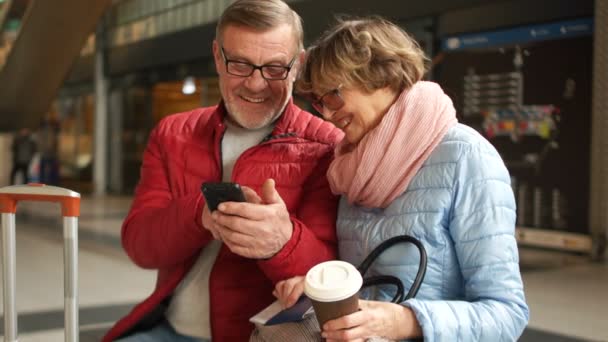 Hombre de barba gris maduro muestra a su esposa una foto de su teléfono inteligente. Ambos se divierten riendo y discutiéndolo. Felices viajes en familia, estación de tren — Vídeos de Stock