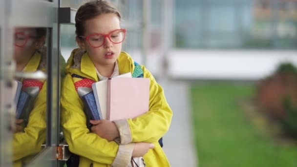 Chica cansada con gafas rojas y una chaqueta amarilla suspira de pie cerca de la escuela. Carga de estudio, dificultades en la escuela, acoso escolar y acoso entre adolescentes — Vídeo de stock