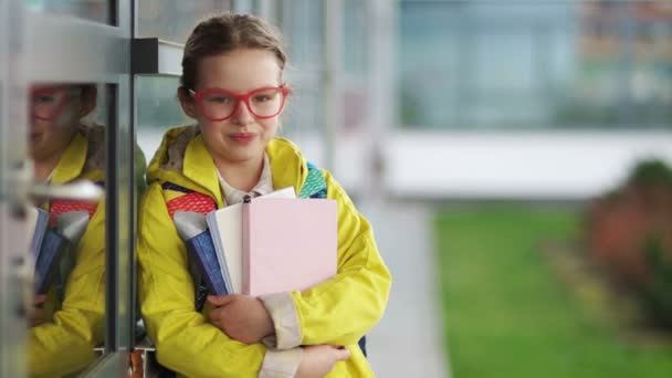 Hermosa chica con gafas y una chaqueta amarilla sosteniendo libros y sonrisas. Colegiala cerca de la escuela, primer día escolar, vacaciones escolares — Vídeo de stock