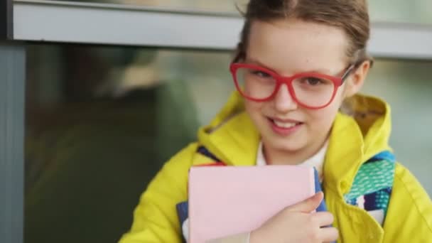 Tioåriga Schoolgirl glatt leende på kameran. Flickan i röda glas och en gul jacka med läro böcker i sina händer — Stockvideo