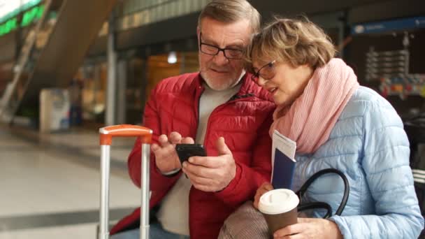 Elegante pareja jubilada en la estación de tren hojeando fotos en su teléfono móvil. Feliz viaje. — Vídeo de stock