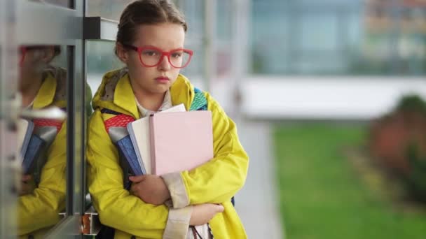 Dulce niña triste de diez años mira a la cámara y luego sonríe. Una colegiala con libros de texto en gafas y una chaqueta amarilla está de pie cerca de la escuela. Regreso a la escuela — Vídeo de stock