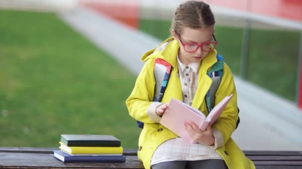 Відмінний студент в жовтій куртці і окулярах сидить на лавці біля школи і читає книгу. Назад до школи, шкільна перерва — стокове відео