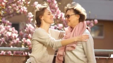 Yetişkin kızı bir çiçeklenme manolya fon karşı balkonda annesi sarılma. Kadınlar bej giysiler giymiş, rüzgar saç geliştirir, parlak bahar güneşi, anneler günü