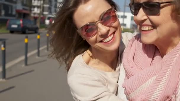 Две женщины, портрет рядом, мать и дочь на городской улице. Солнечный день, женщины в очках смеются и обнимаются — стоковое видео