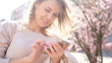 Parlak gökyüzü ve çiçek açan manolya ağaçları arka plan onu akıllı telefon kullanarak büyüleyici gülümseme ile Mutlu kız. Bahar konsepti