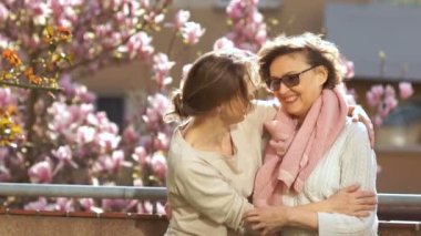 Olgun anne ve yetişkin kızı manolya çiçekli ağacının bir arka plan üzerinde bir balkonda ayakta sarılma. Parlak bahar güneşi, Anneler Günü