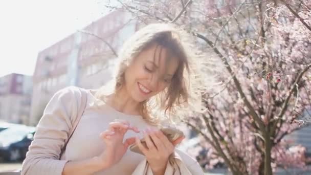 Mulher brilhante e alegre na rua da cidade da primavera com um smartphone na mão. Retrato fechado — Vídeo de Stock