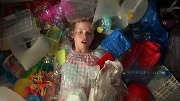 Flickan ligger i en hög med flerfärgade sopor, en plastpåse faller ovanpå henne. Problemet med plast förorening av planeten — Stockvideo
