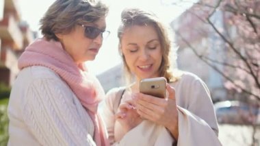 Anne ve yetişkin kızı güneşli rüzgarlı havalarda bir bahar sokak ortasında duran bir akıllı telefon fotoğrafları izliyor. İki kadın ve bir akıllı telefon, anneler günü