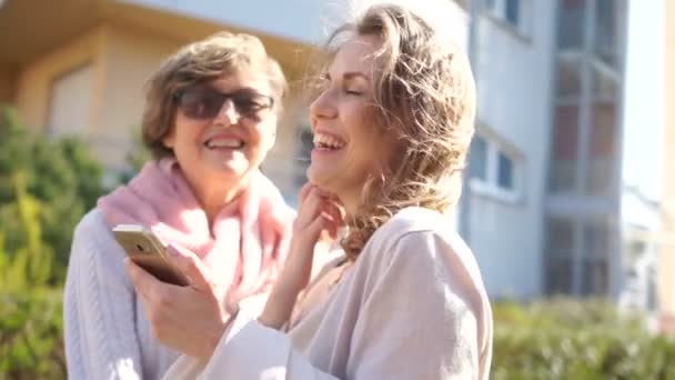 Un retrato alegre de dos mujeres con un smartphone en sus manos. Madre e hija riendo de pie en medio de la calle de primavera sobre el fondo de los árboles florecientes — Vídeo de stock