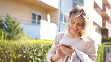 Şehir caddesinde genç ve mutlu bir bahar kadını akıllı telefonuna bakıyor ve gülümsüyor. Parlak güneşli bir sabah