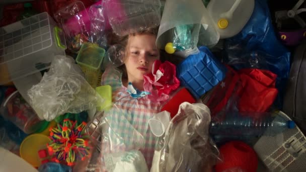 Een kind schoolmeisje is liggend in een stapel van multi-gekleurde vuilnis, een plastic zak valt op de top van haar. Het probleem van plastic vervuiling van de planeet — Stockvideo