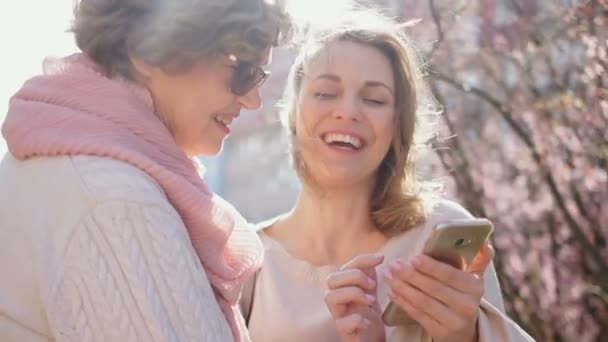 İki kadın akıllı telefondaki bir fotoğrafa bakıyor ve neşeyle gülüyor. Anne ve kızı bahar güneşli rüzgarlı havalarda sokakta dururken konuşuyor — Stok video