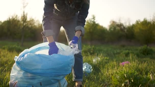 Женщина-волонтер собирает пластиковый мусор в парке. Девушка одета в джинсовый костюм и носит голубые защитные перчатки. Солнце и ветреная погода — стоковое видео