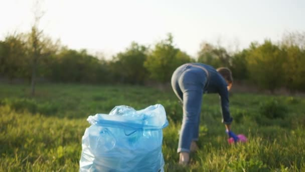 Detener el plástico, concepto social. La chica en jeans recoge basura en el parque en una gran bolsa de plástico azul — Vídeo de stock