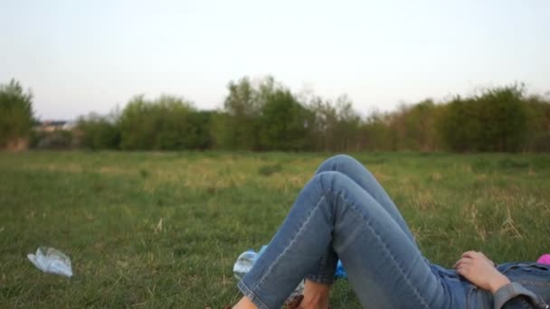 Портрет девушки в джинсовом костюме, лежащей посреди пластикового мусора на траве в парке — стоковое видео