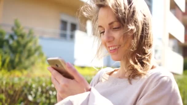 Hermosa chica con un teléfono inteligente en las manos endereza su cabello, de pie en una calle de la ciudad, el sol de primavera brilla intensamente — Vídeos de Stock