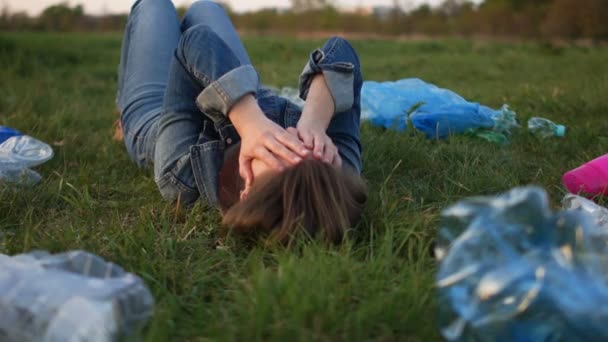 Dziewczyna w garniturze denim leży na trawie wśród plastikowych śmieci. Pokryła jej twarz rękami, kamera wznosi się. Stop tworzywo sztuczne — Wideo stockowe