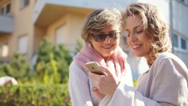 Yetişkin kızı ve onun orta yaşlı annesi güneşli bahar rüzgarlı havalarda caddenin ortasında duran akıllı telefon fotoğrafları izliyor. İki kadın ve bir akıllı telefon, anneler günü — Stok video