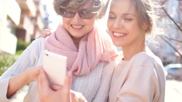 Allegro madre e figlia stanno guardando le foto nello smartphone. Le donne ridono e discutono le loro impressioni. Tempo primaverile, gli alberi sbocciano — Video Stock