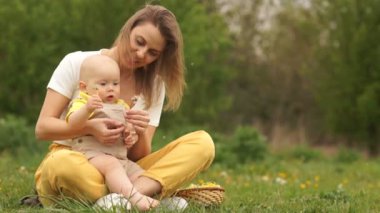 Parkta küçük bir bebeği olan genç bir anne. Çocuk elinde karahindiba tutuyor. Sarı-yeşil renk şeması. Anneler Günü