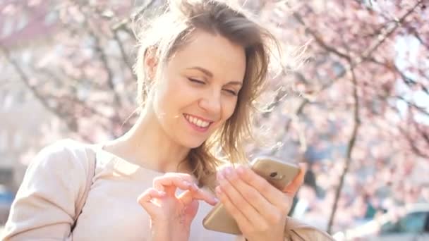 Hermosa joven sosteniendo un teléfono inteligente en sus manos. Retrato exterior, sobre el fondo de árboles de primavera florecientes y sol de primavera brillante — Vídeos de Stock